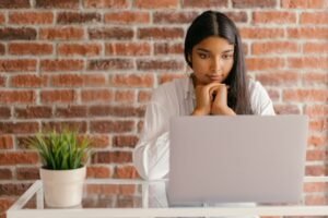 Young woman deeply focused on her laptop studying indoors against a brick wall backdrop.