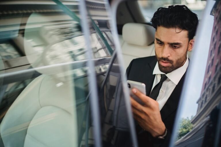Confident businessman in suit using smartphone inside car, viewed through window reflection.