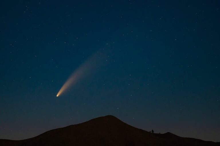 A breathtaking capture of a comet streaking across a star-filled night sky above a mountain silhouette.