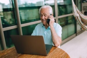 Elderly man in a blue shirt talking on phone while using laptop indoors. Ideal for remote work concepts.