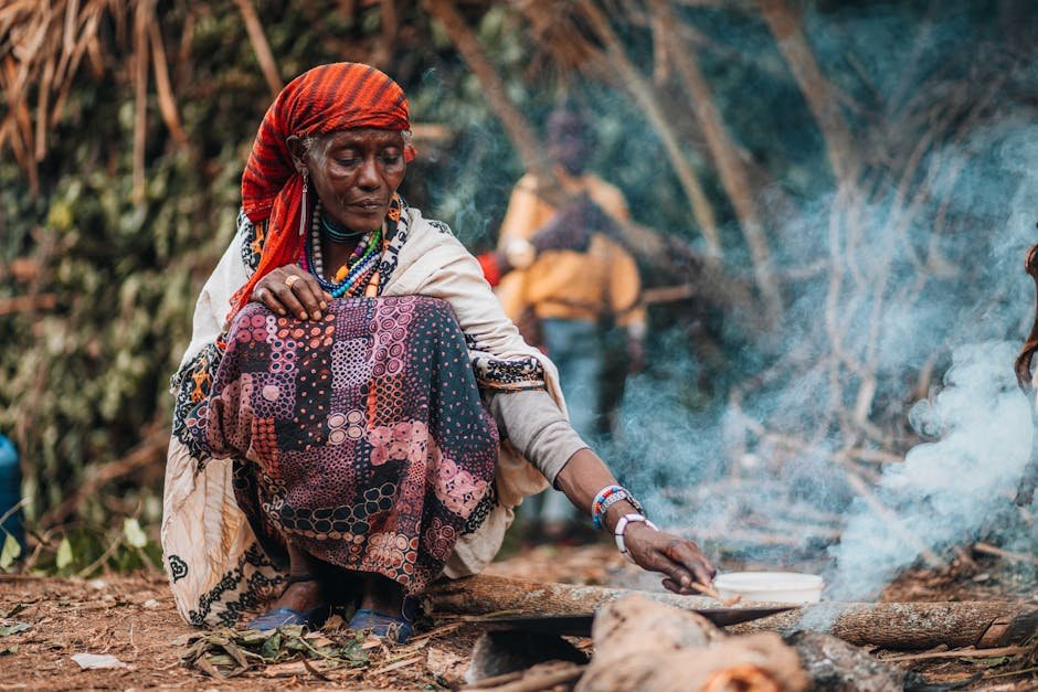 A woman cooking outdoors in a traditional African village setting with visible smoke.