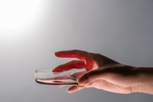 A hand carefully holds a petri dish containing a red liquid, against a gray background.