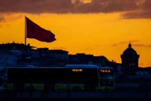 Dramatic urban landscape featuring a city bus and national flag silhouette against a vivid sunset sky.
