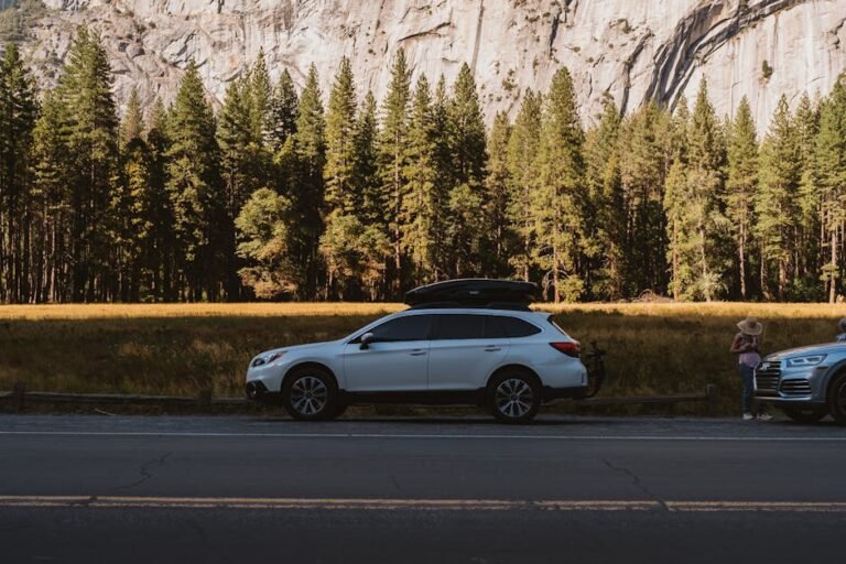 A white SUV parked on a peaceful road beside lush trees and rocky cliffs possibly in Yosemite National Park.