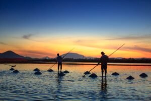 Stunning silhouette of farmers harvesting salt in a tranquil field at sunset, reflecting warm colors on the water.