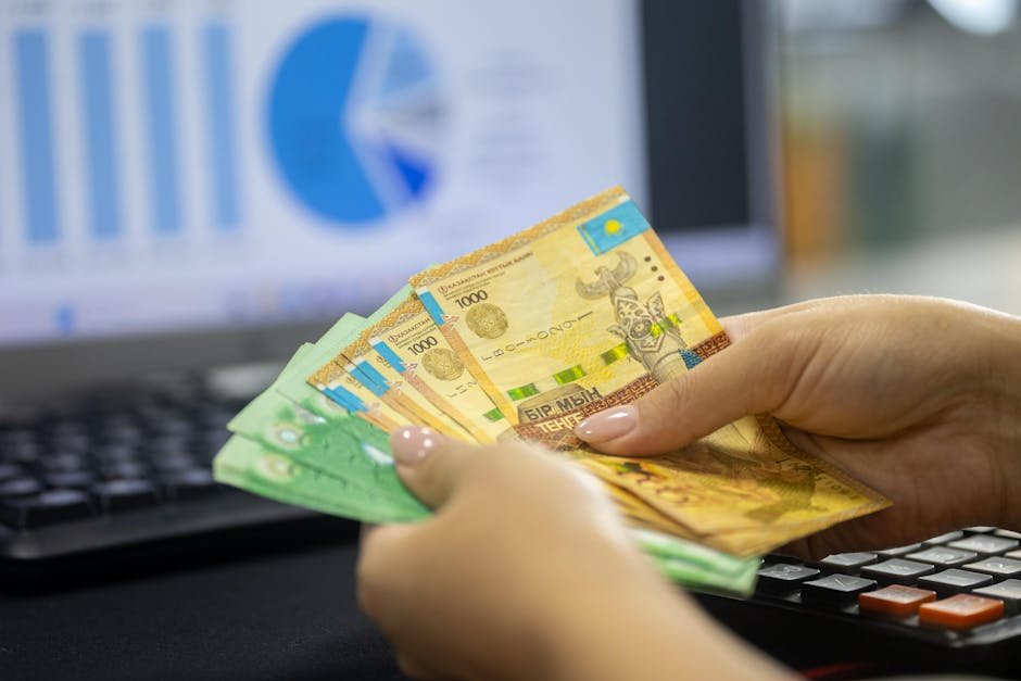 Human hands counting Kazakh tenge bills near a laptop displaying financial graphs.